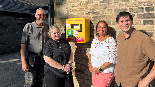 Church Warden Steven Reed, Vicar of Leesfield and Hey Reverend Kirsten Stott, Councillor Alicia Marland, Councillor Sam Al Hamdani at St John's Church with the defibrillator